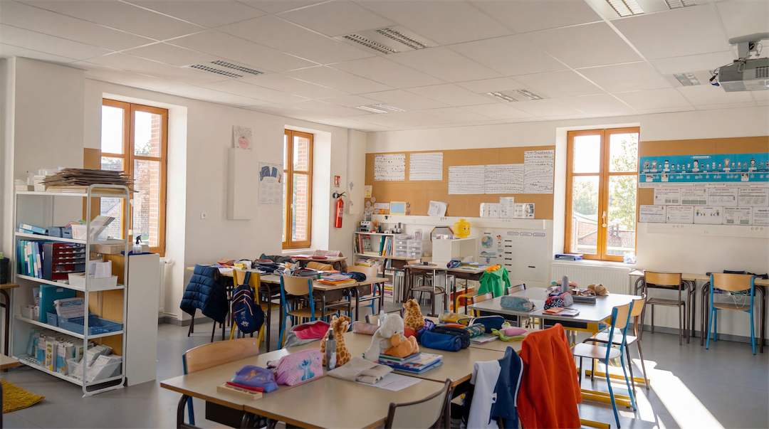 Classe d'école primaire avec tables regroupées, chaises, sacs à dos, peluches et matériel scolaire sous lumière naturelle.