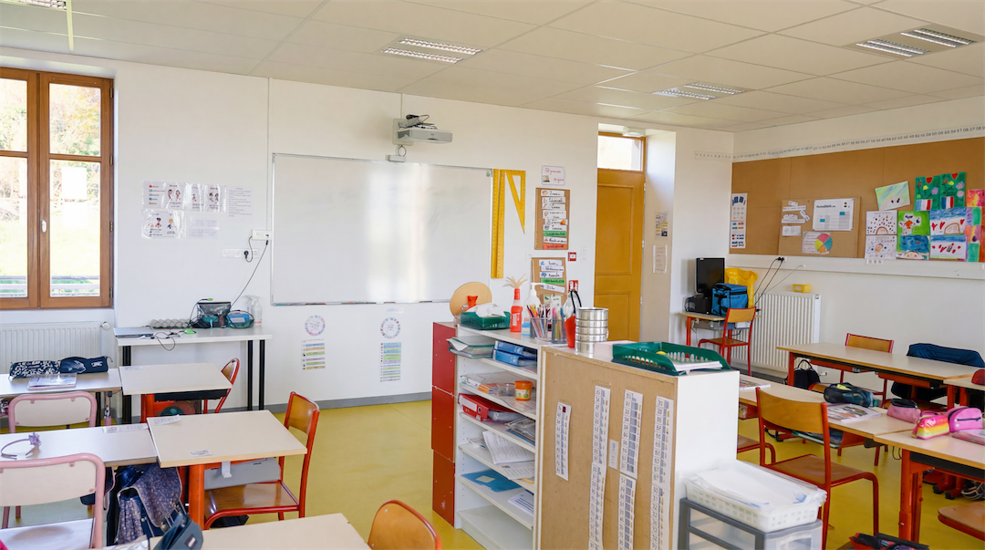 Classe d'école primaire bien éclairée avec bureaux, chaises rouges, tableau blanc, étagères remplies de fournitures et dessins accrochés aux murs.