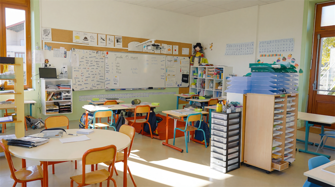 Classe d'école avec tables rondes et rectangulaires, chaises colorées, tableau blanc avec notes et rangements organisés.