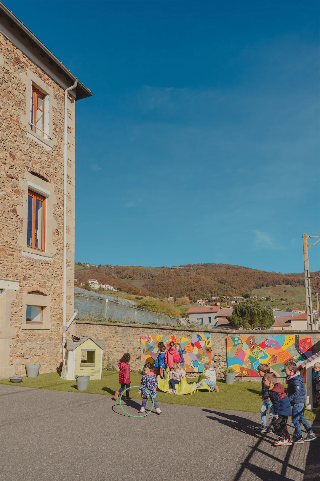 Enfants jouant dans une cour d'école extérieure avec des panneaux muraux colorés et une petite cabane près d'un bâtiment en pierre sous un ciel bleu.