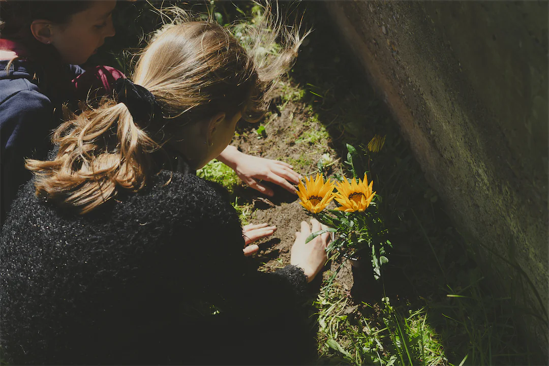 Deux enfants plantent des fleurs jaunes dans le sol d'un jardin ensoleillé près d'un mur.