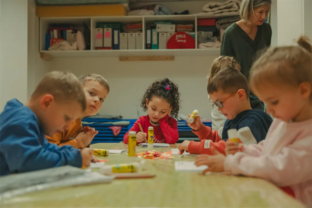 Des enfants assis autour d'une table effectuent une activité créative avec des bâtons de colle, tandis qu'une adulte supervise en arrière-plan.
