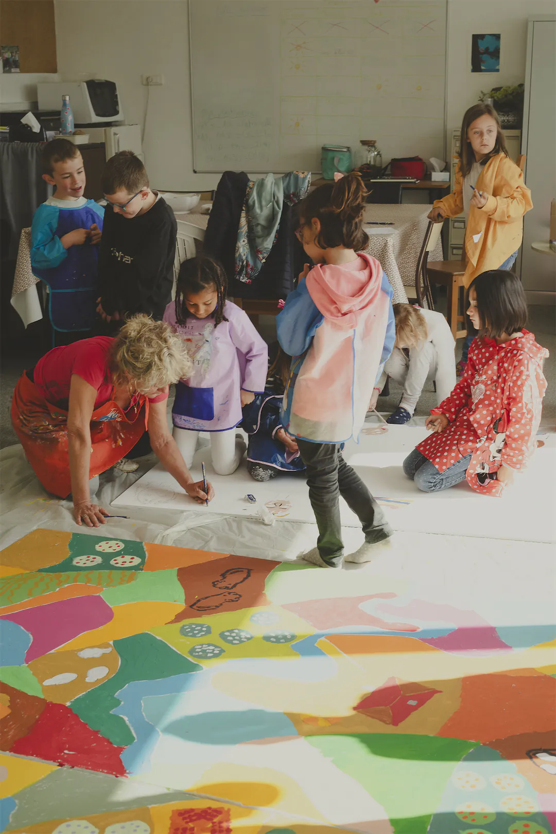 Une enseignante et plusieurs enfants peignent et dessinent ensemble sur une grande toile colorée au sol dans une salle de classe.