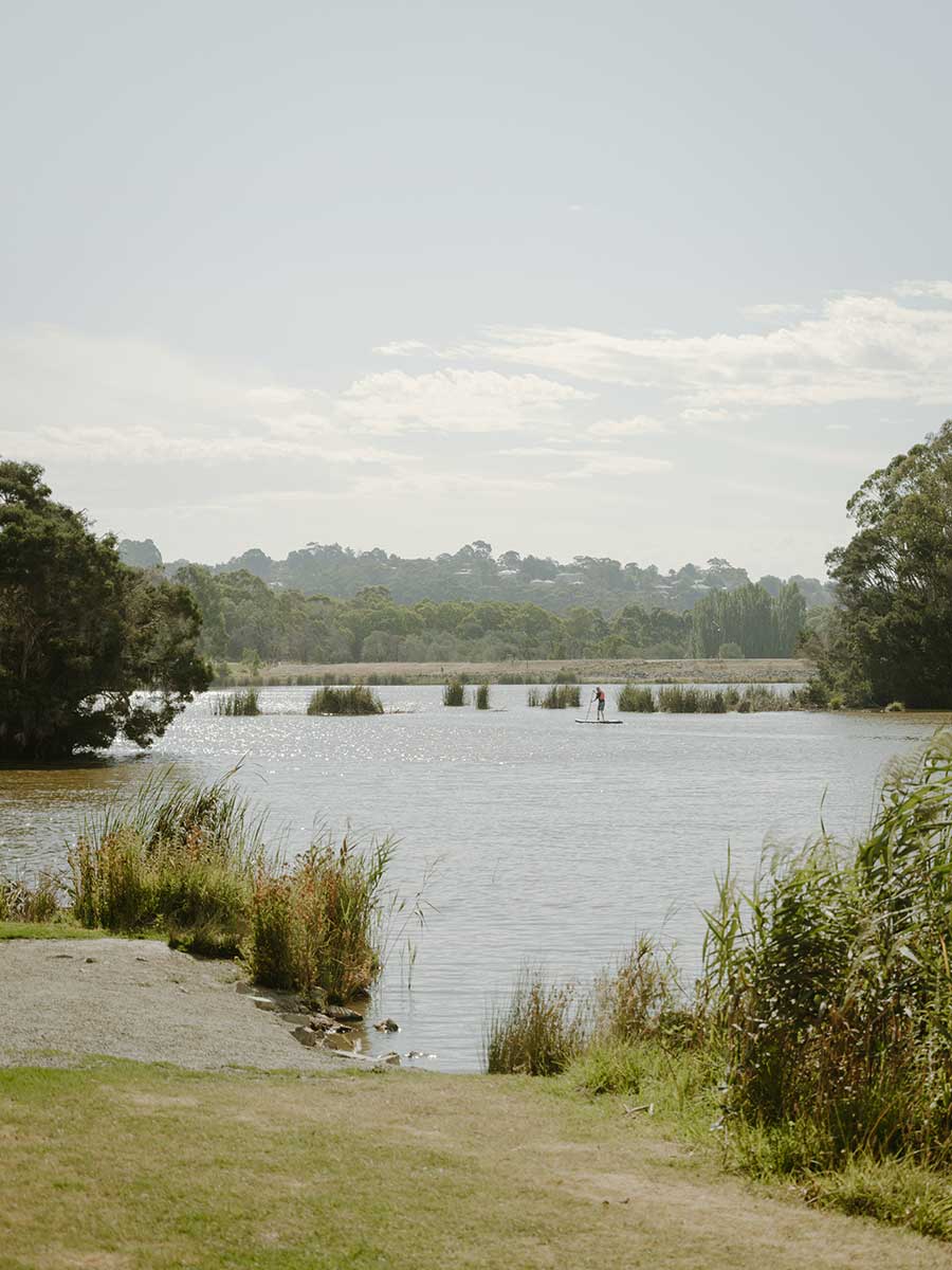 Person paddleboarding on a calm lake surrounded by greenery and trees under a partly cloudy sky.