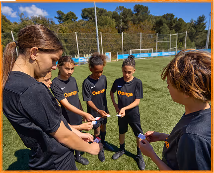 A group of six children in black sports uniforms standing in a circle on a grass field, holding playing cards.