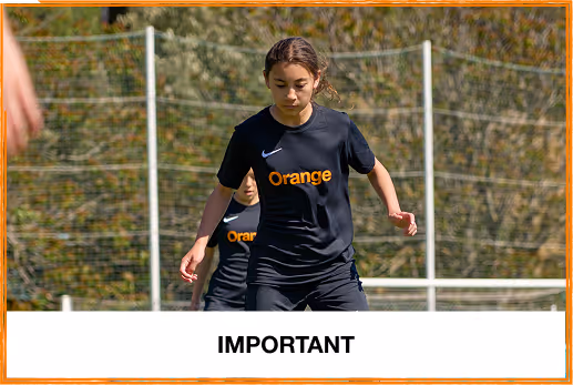 Young female soccer player wearing a black Nike jersey with 'Orange' text, focusing on the ball during practice on a grassy field.