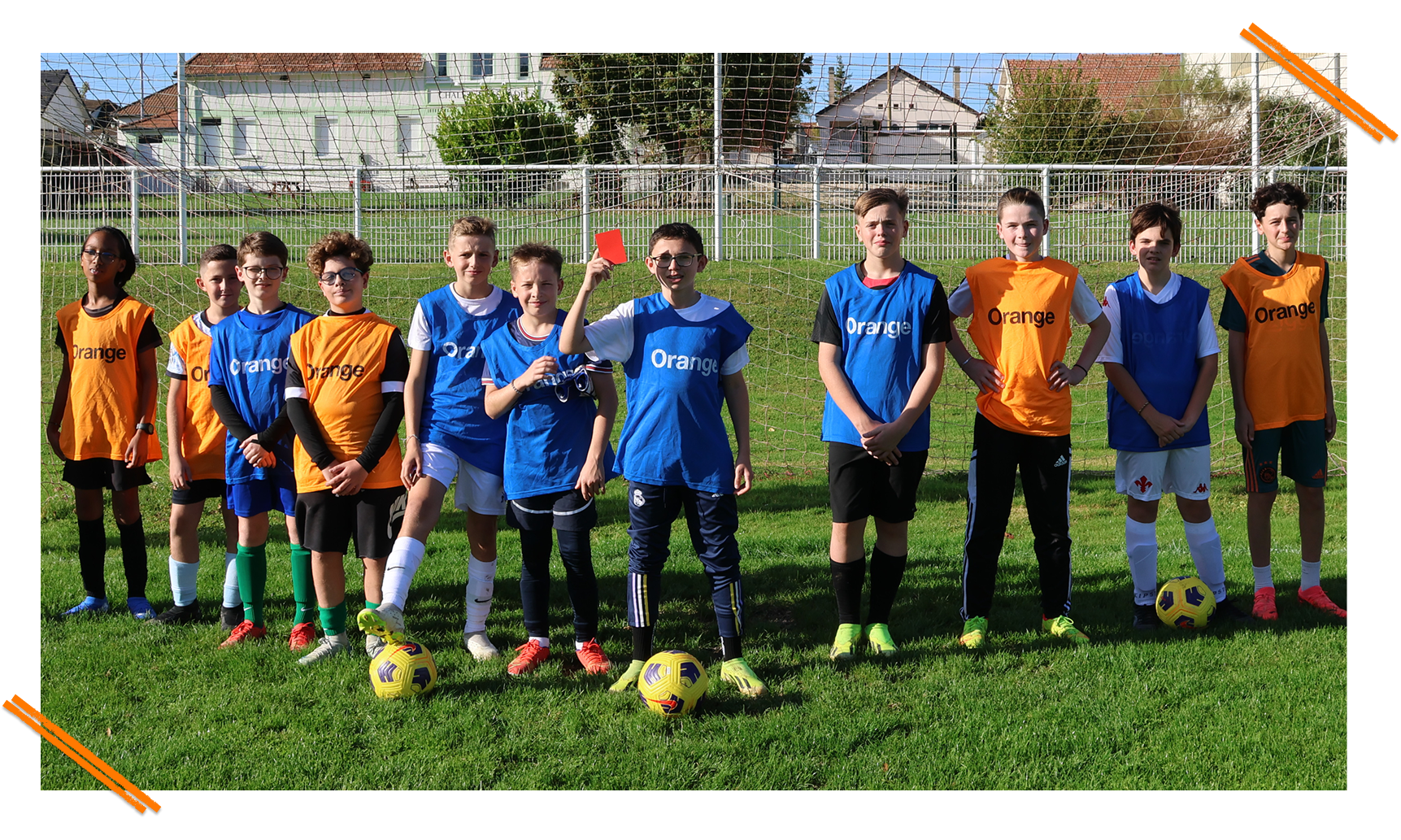 Group of young soccer players in orange and blue jerseys standing on grass in front of a goal, one player holding up a red card.