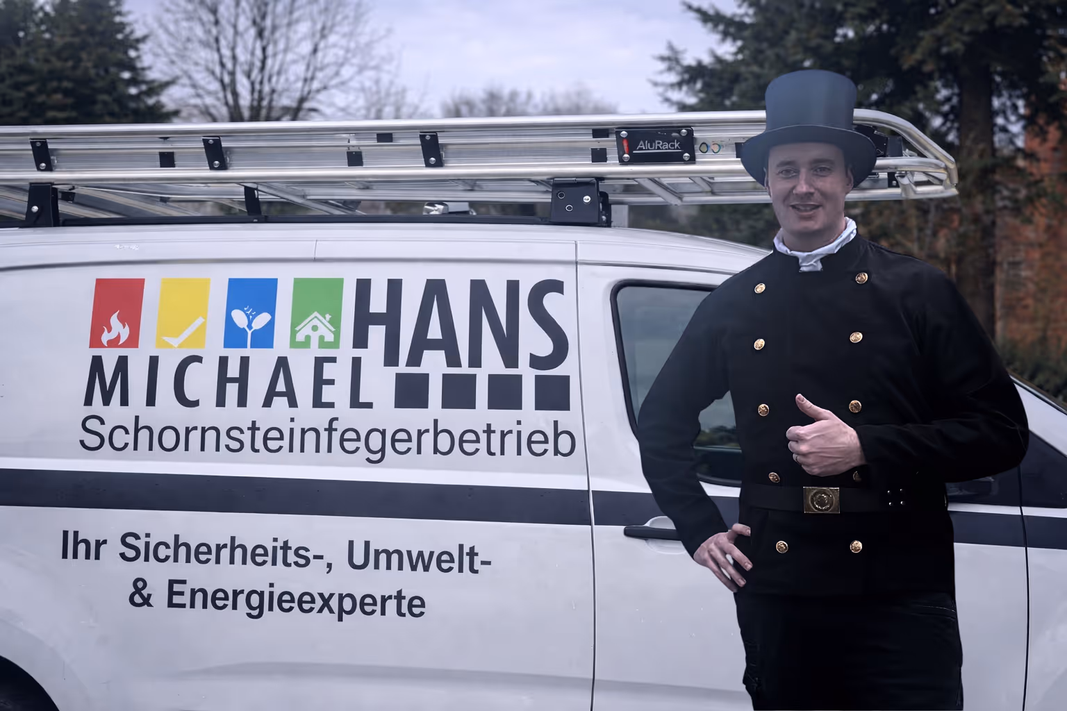 Schornsteinfeger Michael Hans steht rechts vor seinem Firmenwagen mit der Aufschrift Michael Hans Schornsteinfegerbetrieb im Einsatzgebiet Moers und Goch