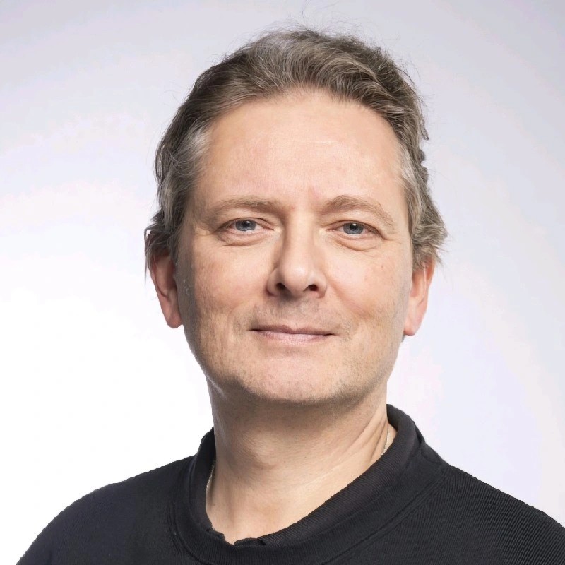 Close-up portrait of a middle-aged man with light skin and blue eyes wearing a black shirt against a light background.
