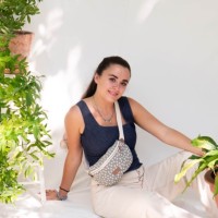 Young woman with long dark hair sitting outdoors among green plants, wearing a navy tank top and white pants with a patterned crossbody bag.