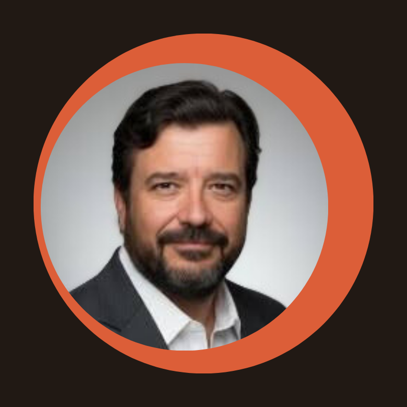 Headshot of a man with dark hair and beard wearing a dark suit jacket and white shirt against a gray background.