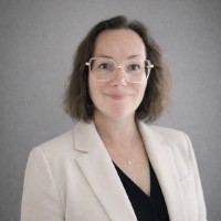 Smiling woman with shoulder-length brown hair wearing glasses, a light beige blazer, and a black top against a gray background.