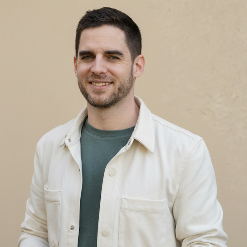 Smiling man with short dark hair and beard wearing a green shirt and cream jacket against a beige background.