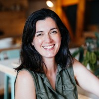 Smiling woman with dark hair wearing a sleeveless green top sitting indoors.