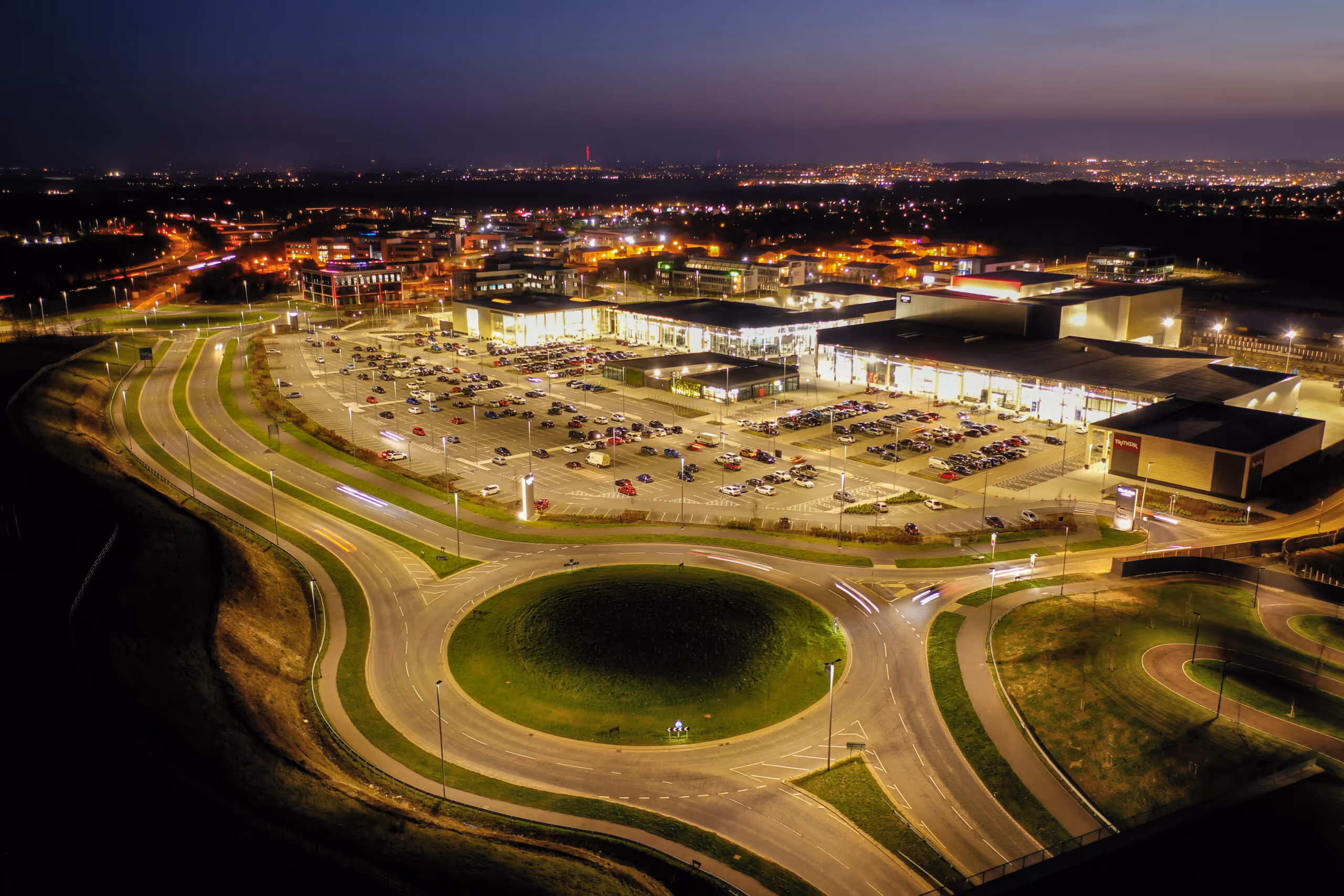 Aerial night view of a brightly lit shopping center with a large parking lot and a roundabout in the foreground.