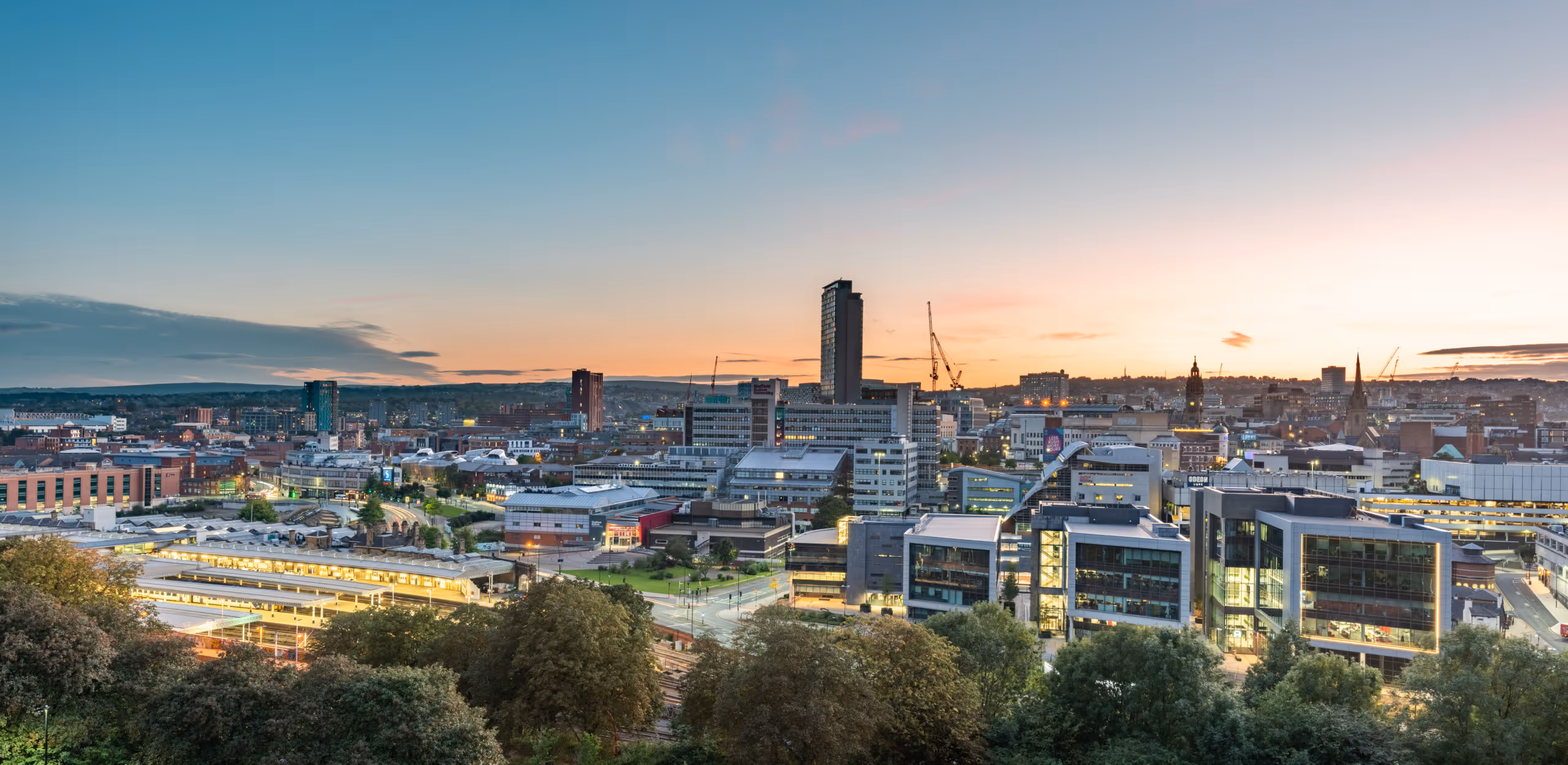 City skyline at sunset with modern buildings, cranes, and green trees in the foreground.