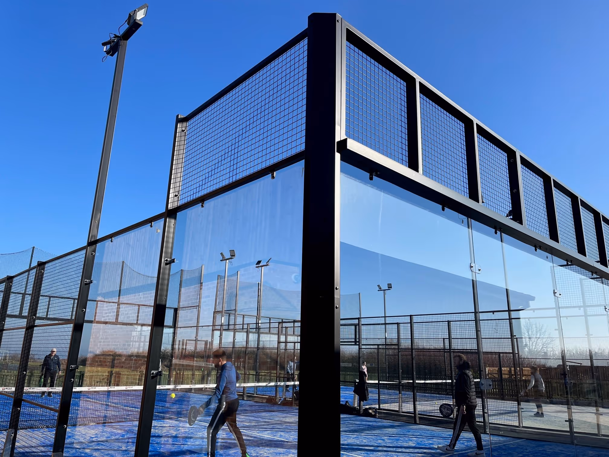 People playing padel on blue outdoor courts surrounded by glass and metal fencing under a clear blue sky.