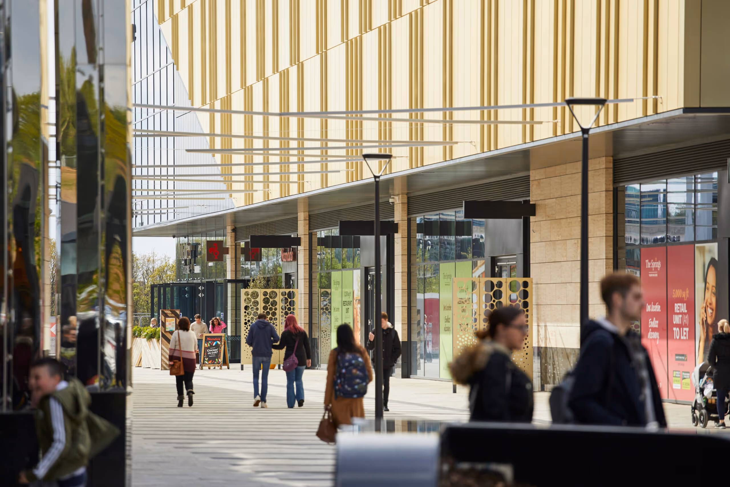 People walking along a modern shopping center with glass storefronts and yellow panelled upper façade.