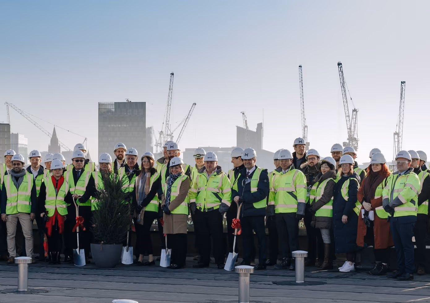Group of construction workers and professionals wearing hard hats and yellow safety vests standing together with city cranes and buildings in the background.
