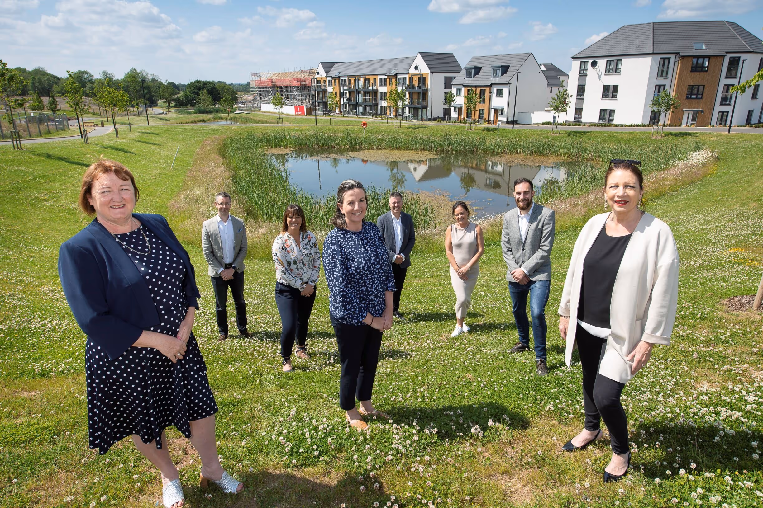 A group of eight people standing spaced out on a grassy area near a small pond with modern residential buildings in the background under a partly cloudy sky.