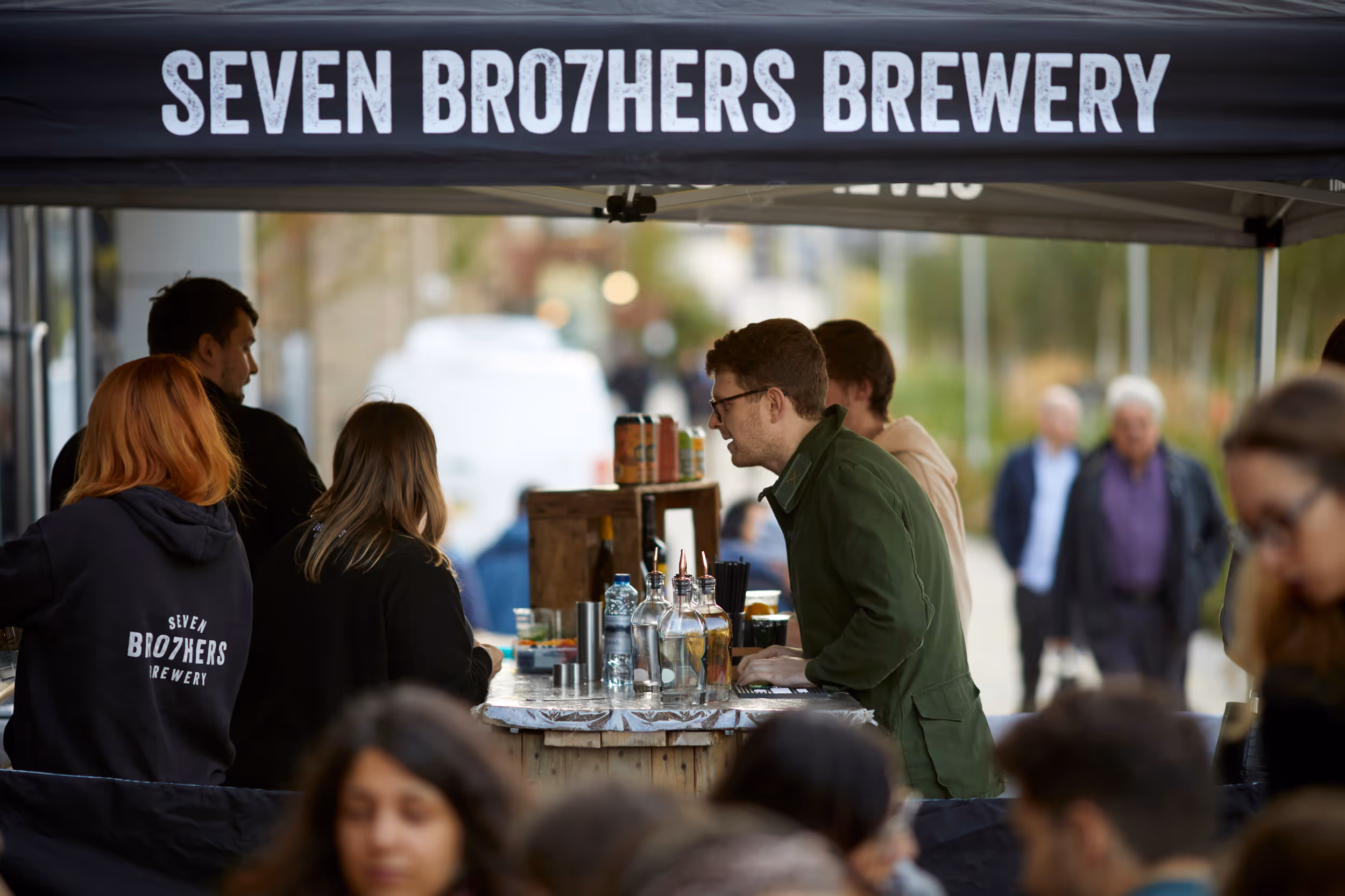 People gathered around a bar under a Seven Bro7hers Brewery tent at an outdoor event.