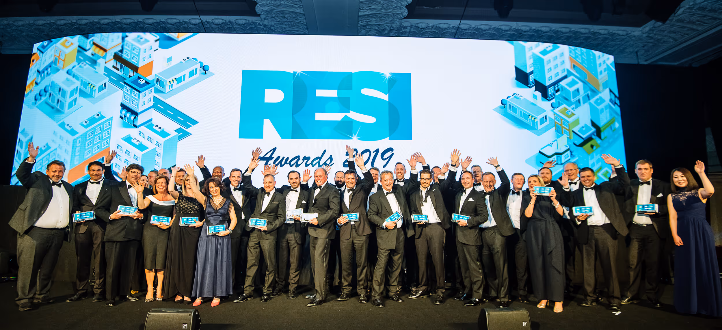 Group of people in formal attire holding RESI Awards trophies and waving on stage in front of a large RESI Awards 2019 screen.