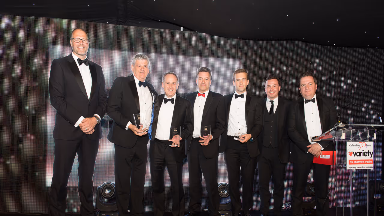 Seven men in black tuxedos standing on stage at an awards event, some holding glass trophies.