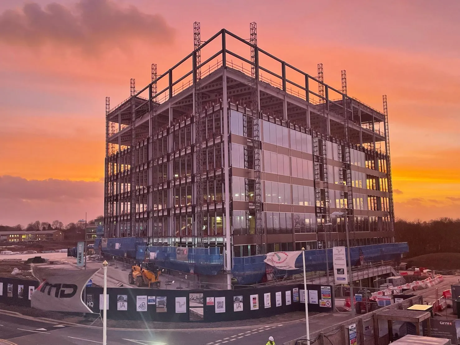 Modern multi-story building under construction at sunset with steel framework and glass panels reflecting the orange sky.
