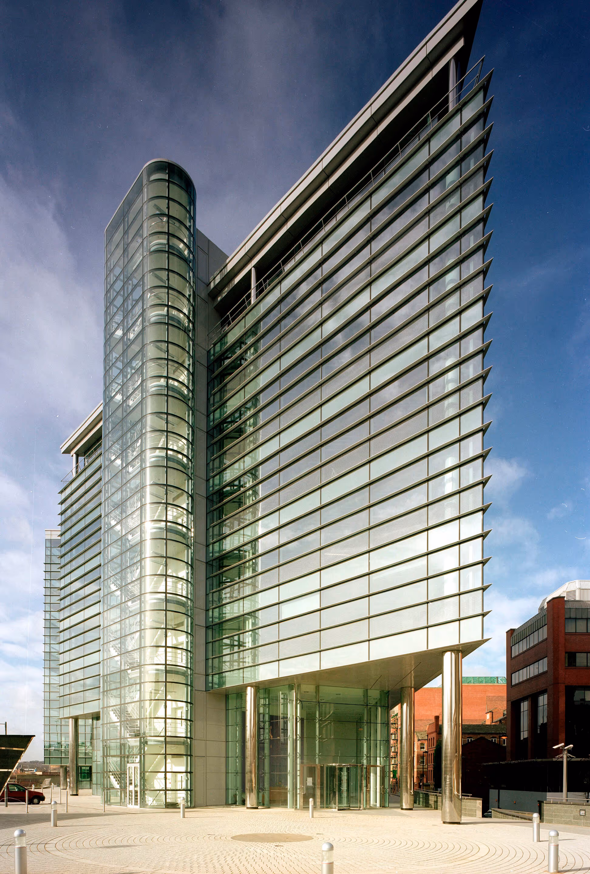 Modern glass office building with a cylindrical glass staircase and reflective windows under a partly cloudy sky.