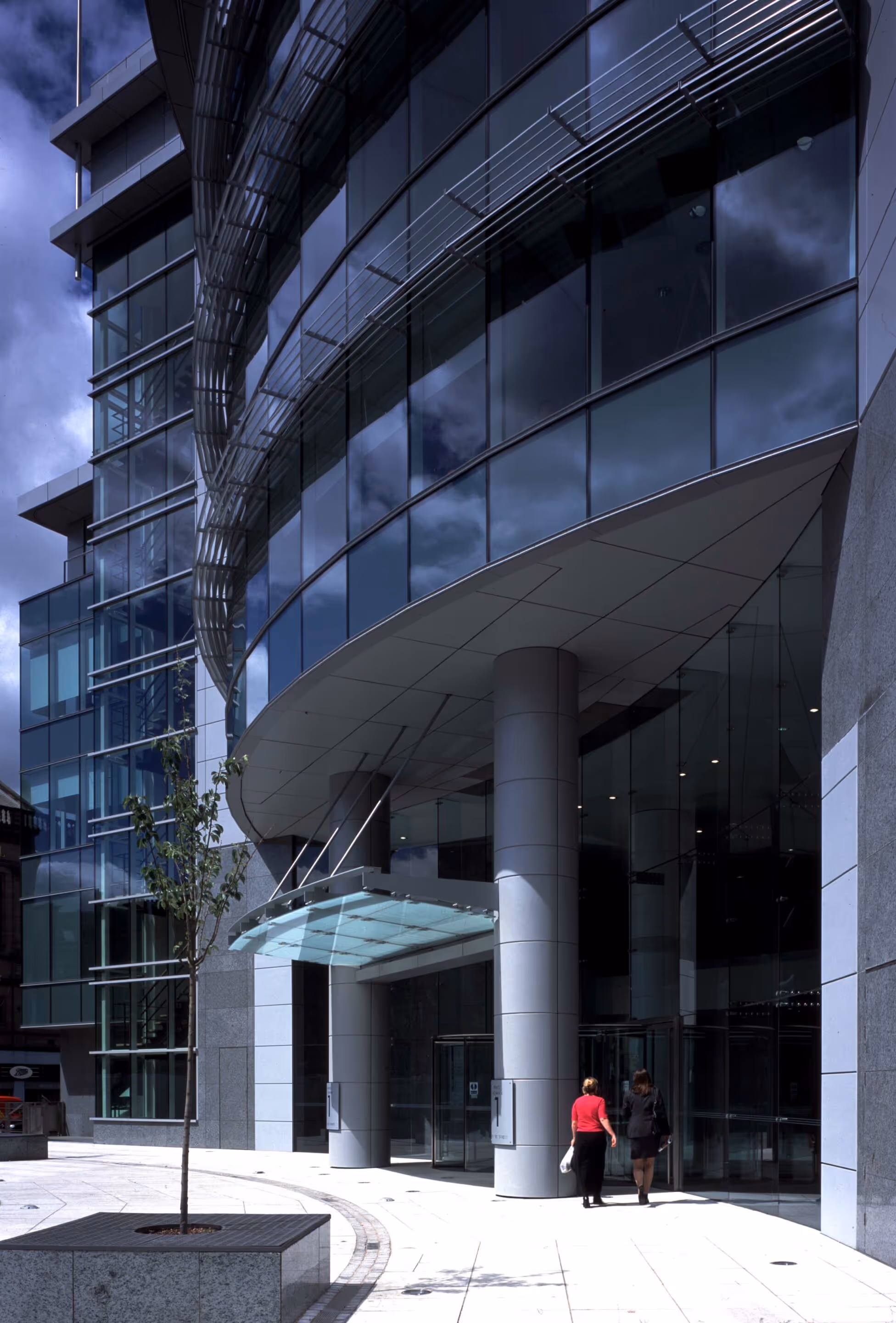 Modern office building entrance with glass facade, curved canopy, and two people walking inside.