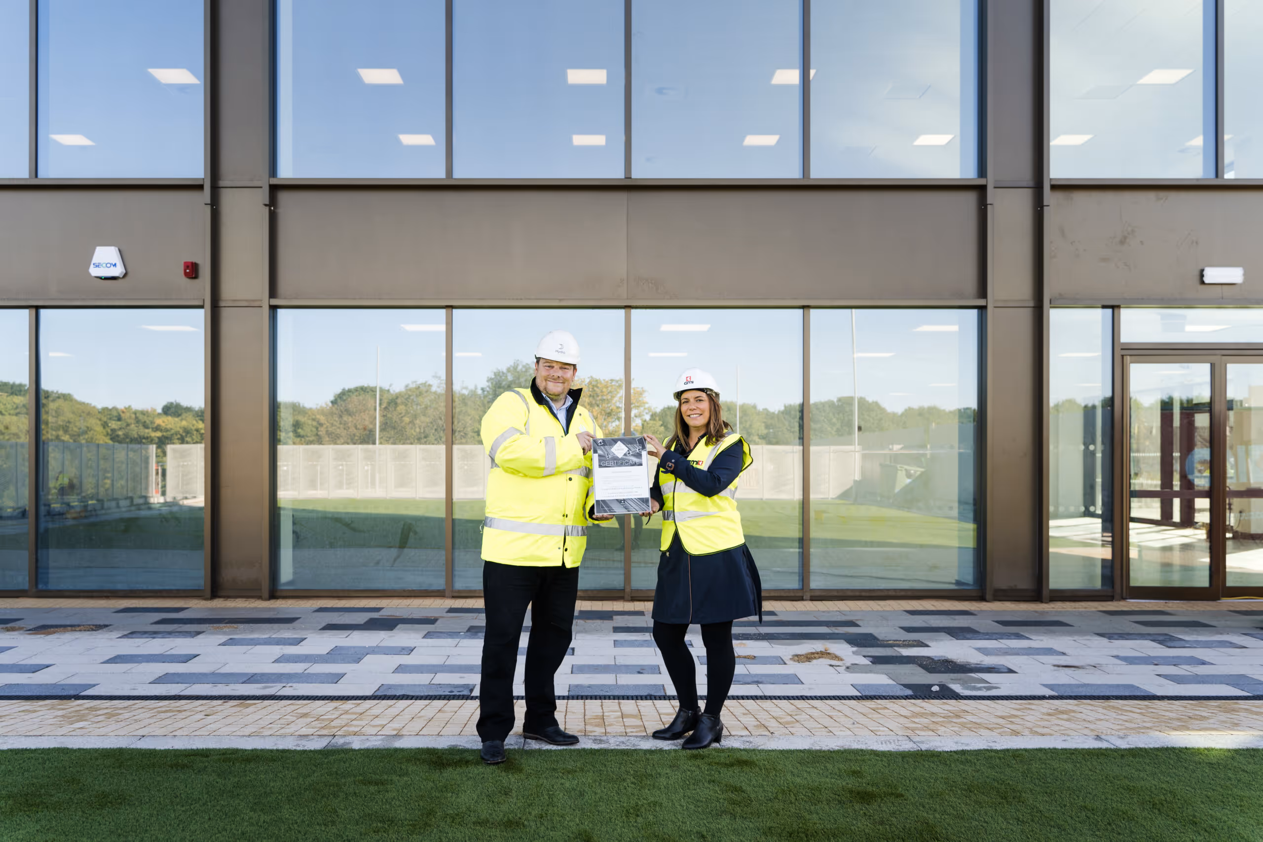 Two construction workers wearing yellow safety vests and helmets holding a certificate in front of a modern building.