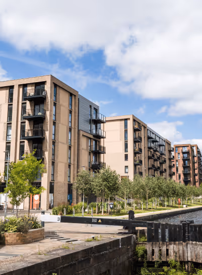 Modern apartment buildings alongside a canal with a lock, surrounded by greenery under a partly cloudy sky.