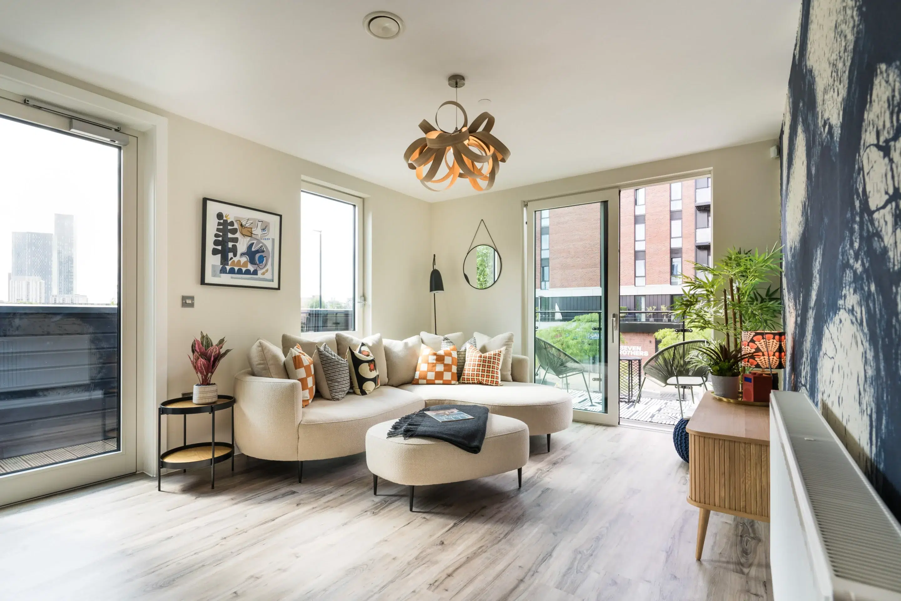Bright living room with curved beige sofa adorned with patterned cushions, round ottoman, modern light fixture, and large sliding glass doors leading to a balcony.