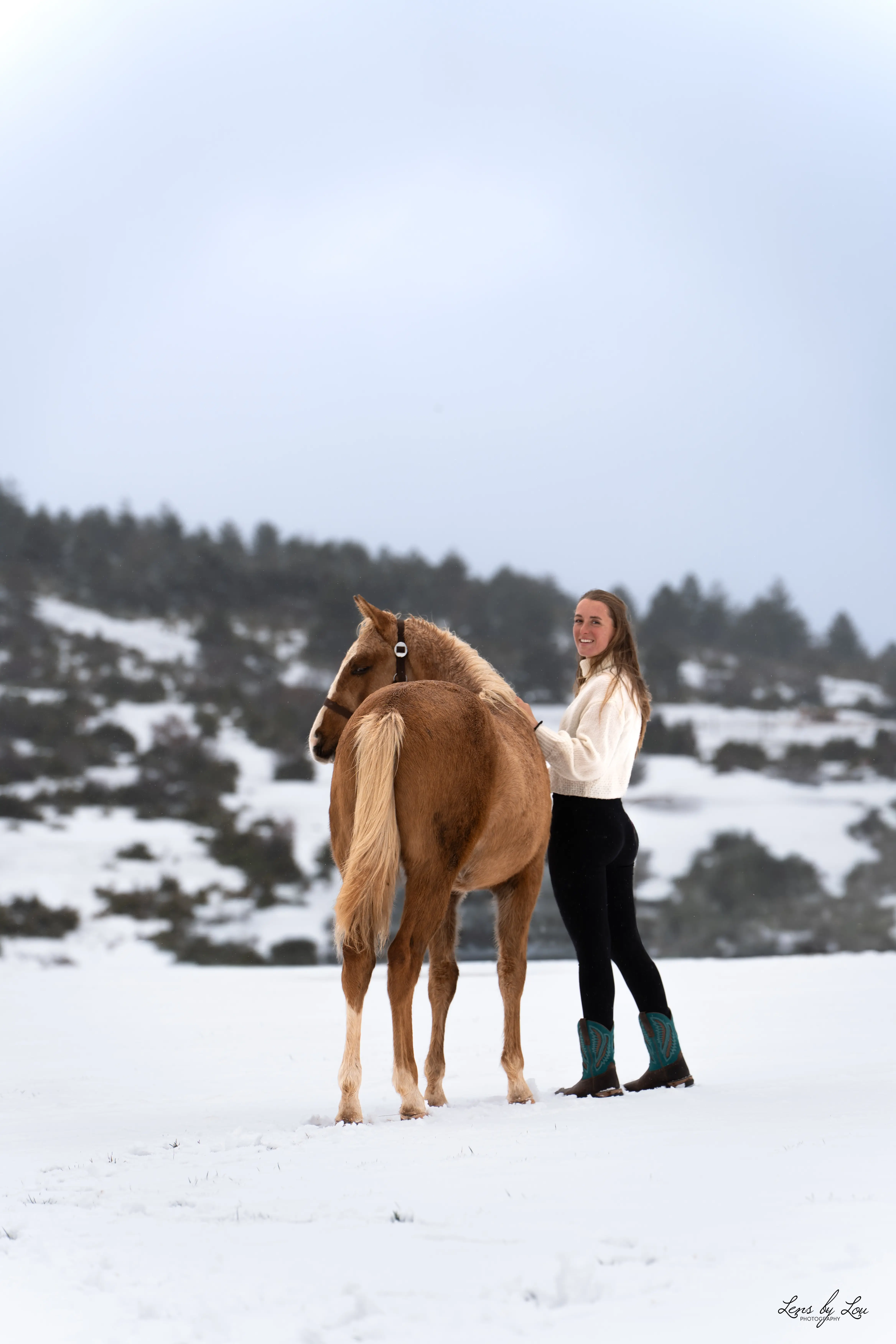 Jeune femme souriante en pull blanc et bottes bleues tenant un cheval alezan dans la neige avec des collines boisées en arrière-plan.