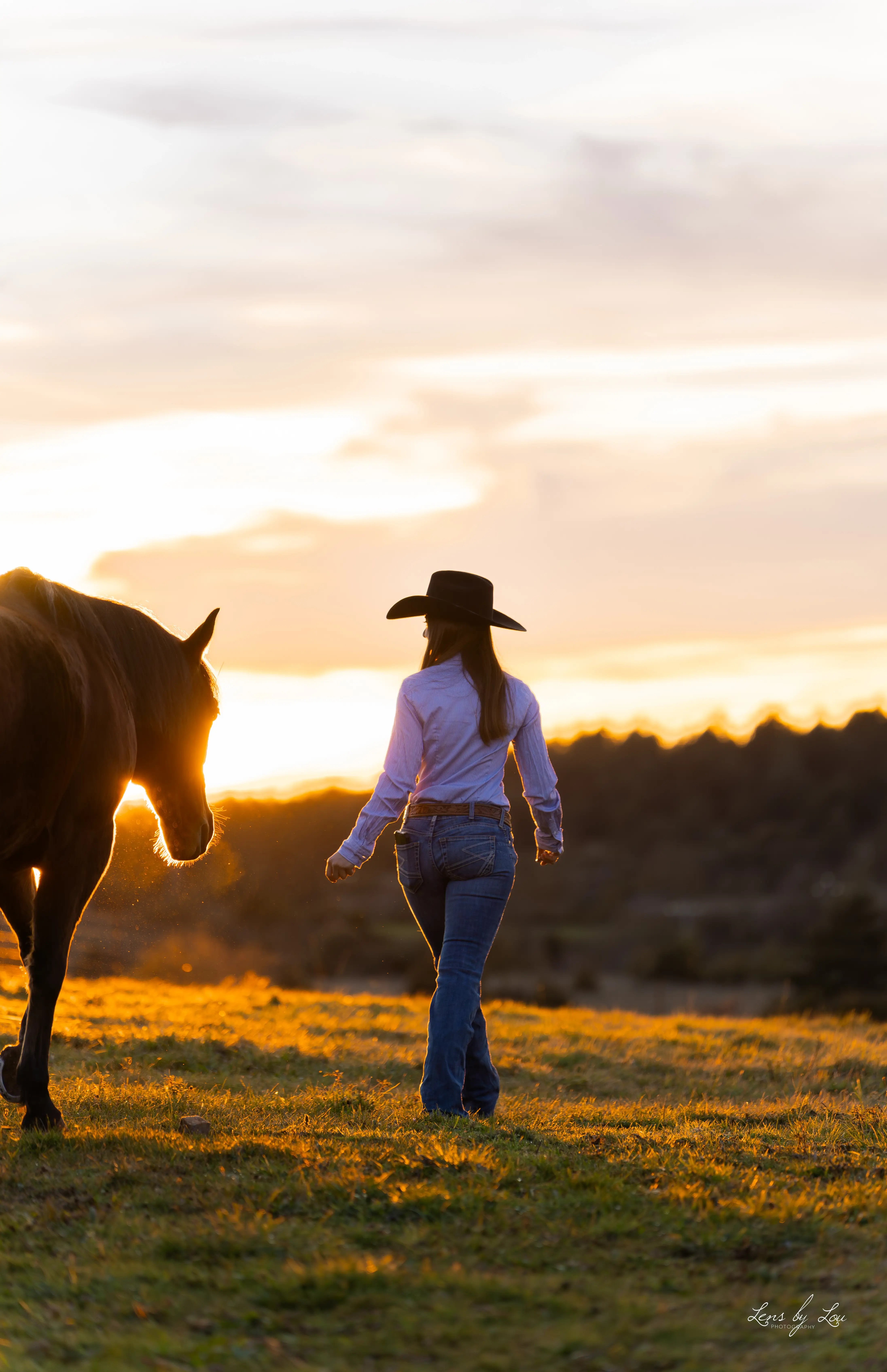 Femme portant un chapeau de cow-boy marchant auprès d'un cheval au coucher du soleil dans un champ.