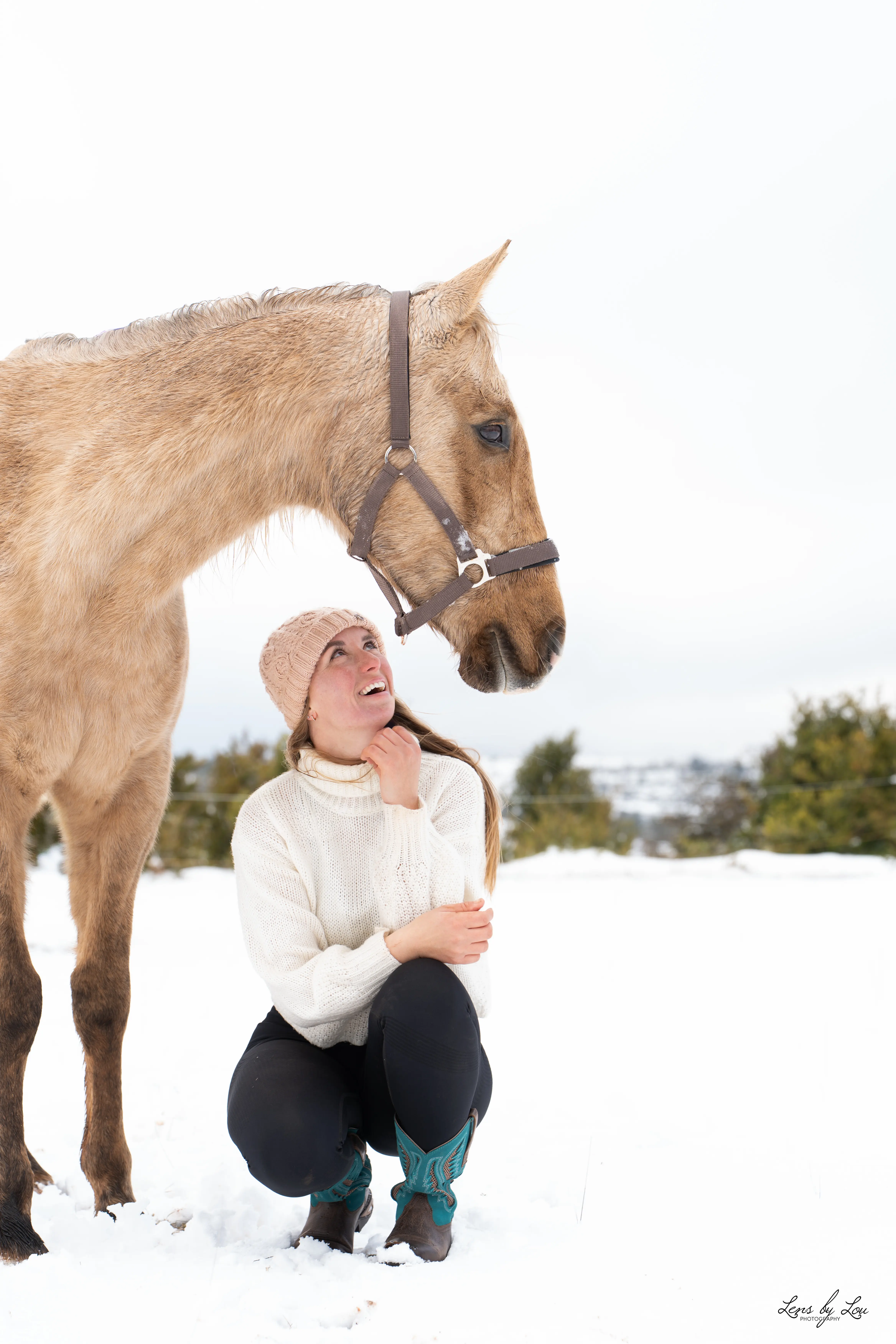 Une femme accroupie dans la neige regarde avec affection un cheval beige à côté d'elle.