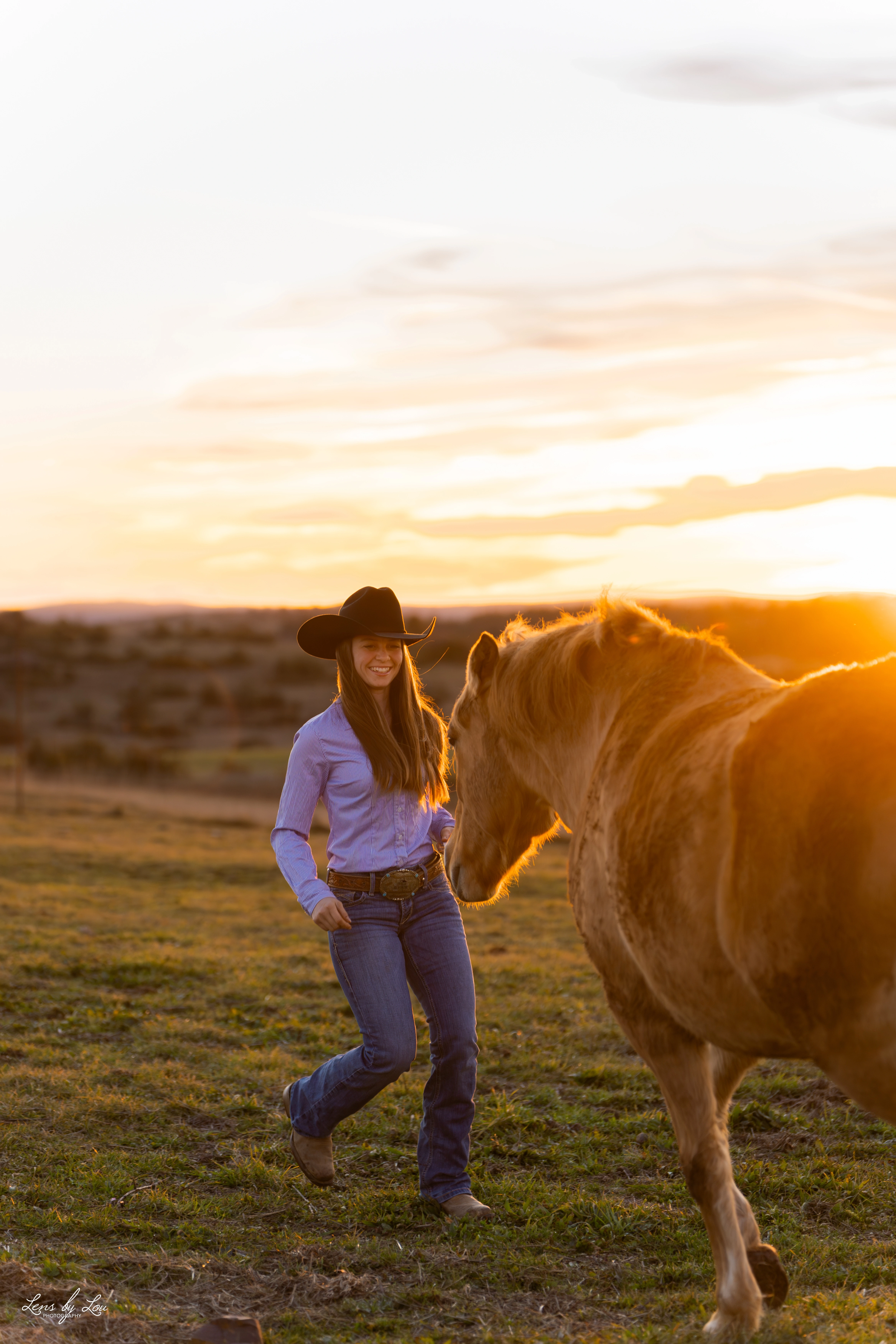 Jeune femme souriante portant un chapeau de cow-boy marchant vers un cheval au coucher du soleil dans un champ.