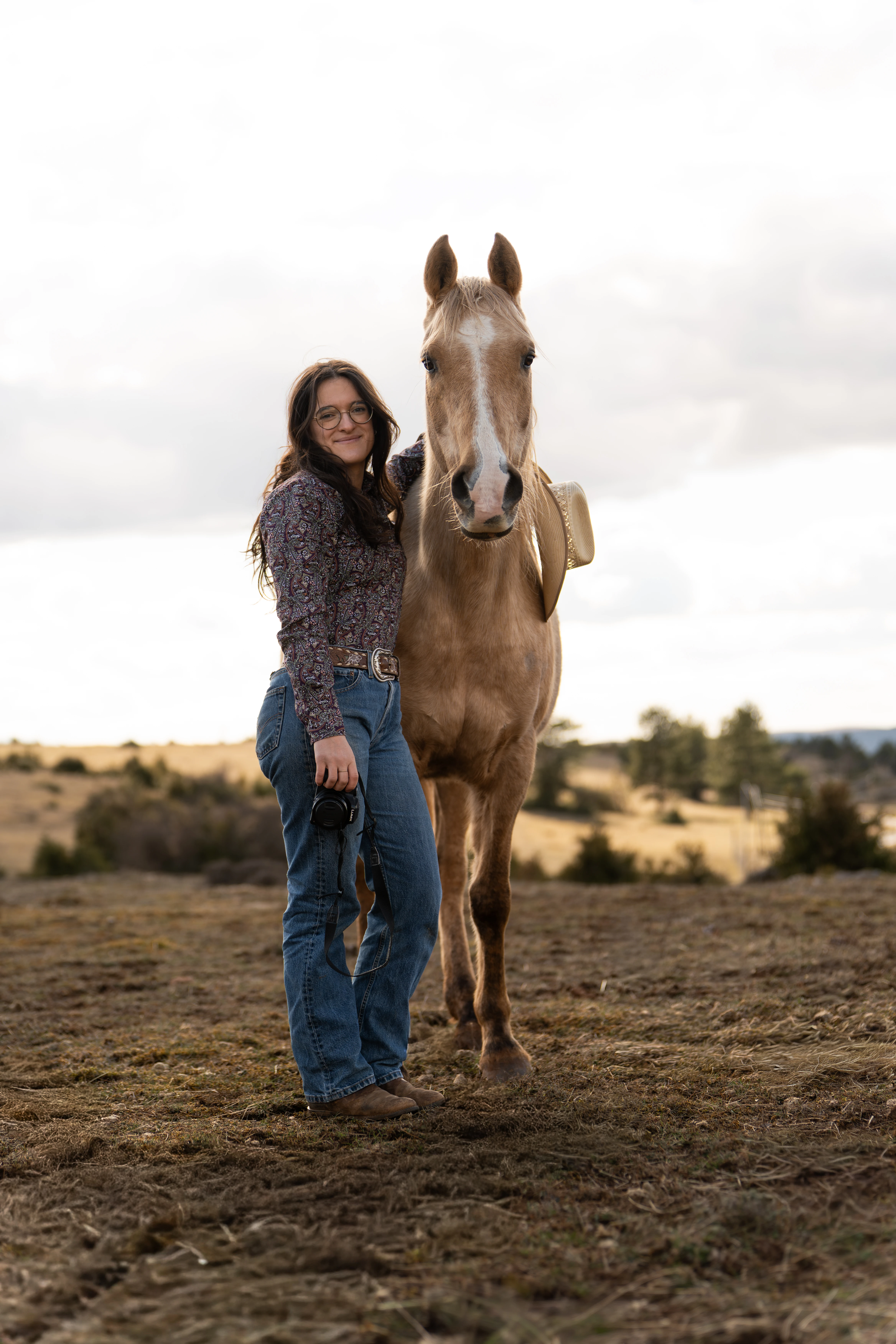Femme souriante avec des lunettes, tenant un appareil photo, posant avec un cheval beige portant un chapeau dans un champ.