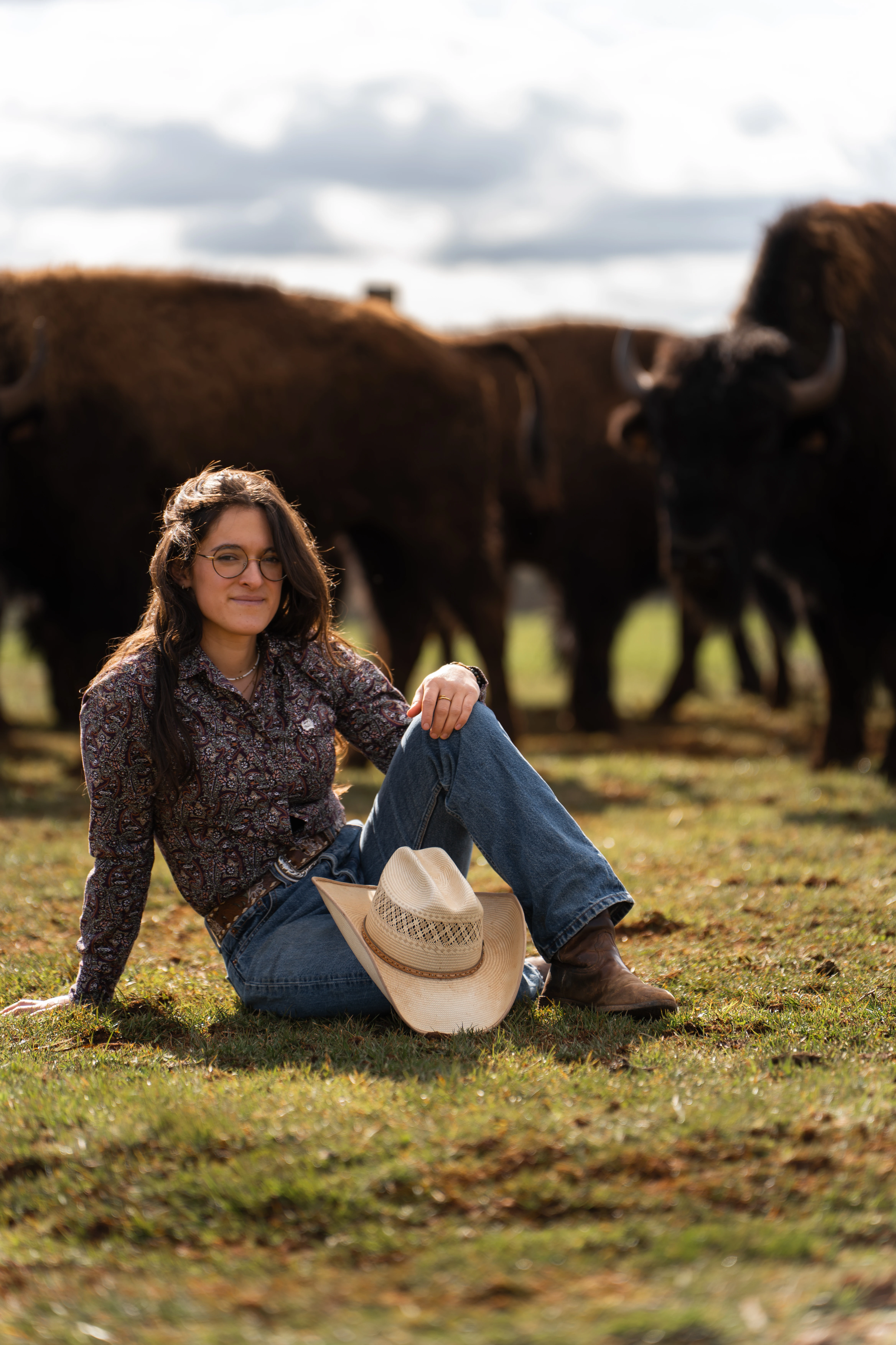 Une femme accroupie en chapeau de cowboy et lunettes avec des bisons dans un champ en arrière-plan.