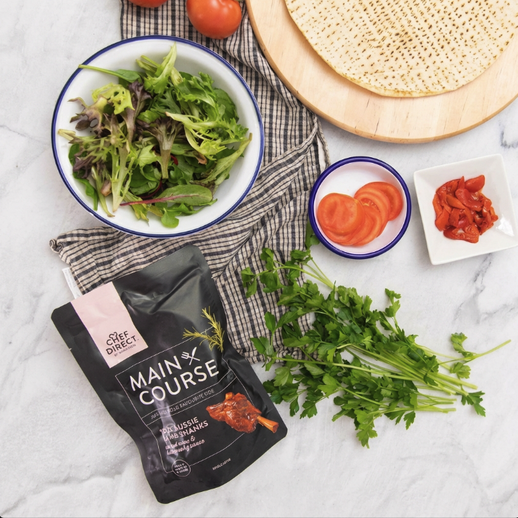 Chef Direct Lamb Shank Pouch with bowl of mixed lettuce leave, a board, herbs, sliced tomato and tea towel