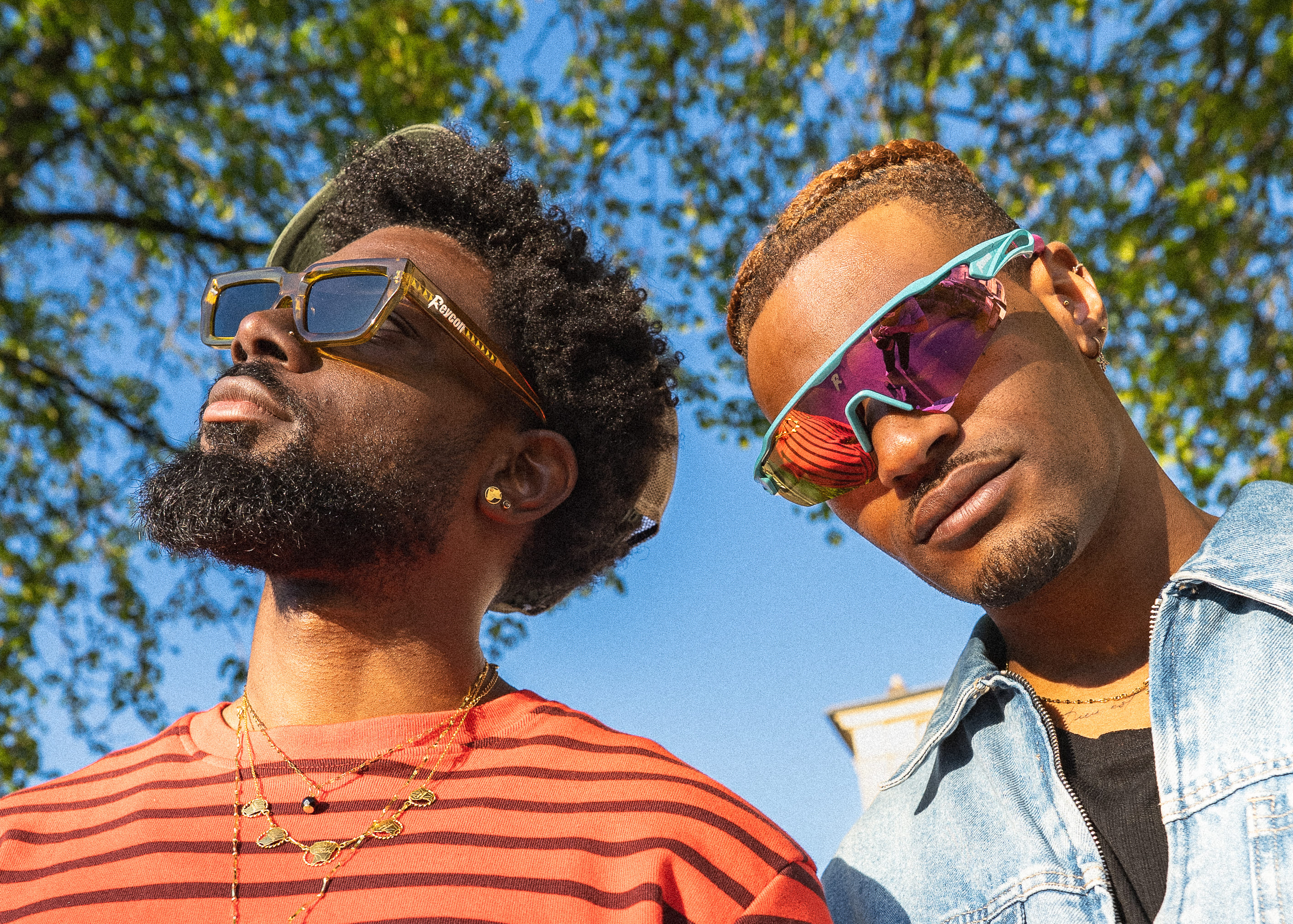 Deux hommes portant des lunettes de soleil, un en rayures rouges et l'autre en veste en jean, sous un ciel bleu.