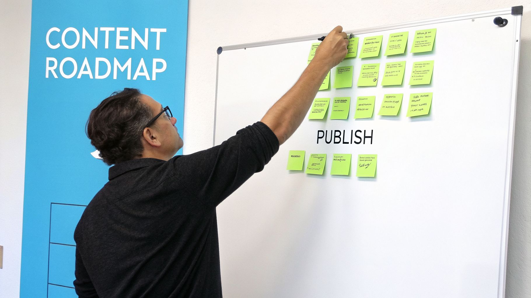 Man with glasses adding content roadmap ideas on green sticky notes to a whiteboard.