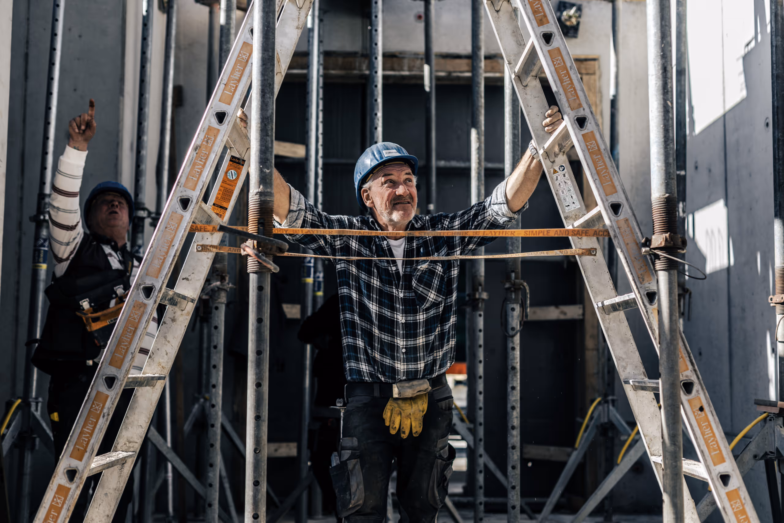 Baumeister mit blauem Helm und kariertem Hemd klettert auf eine Metallleiter auf einer Baustelle.