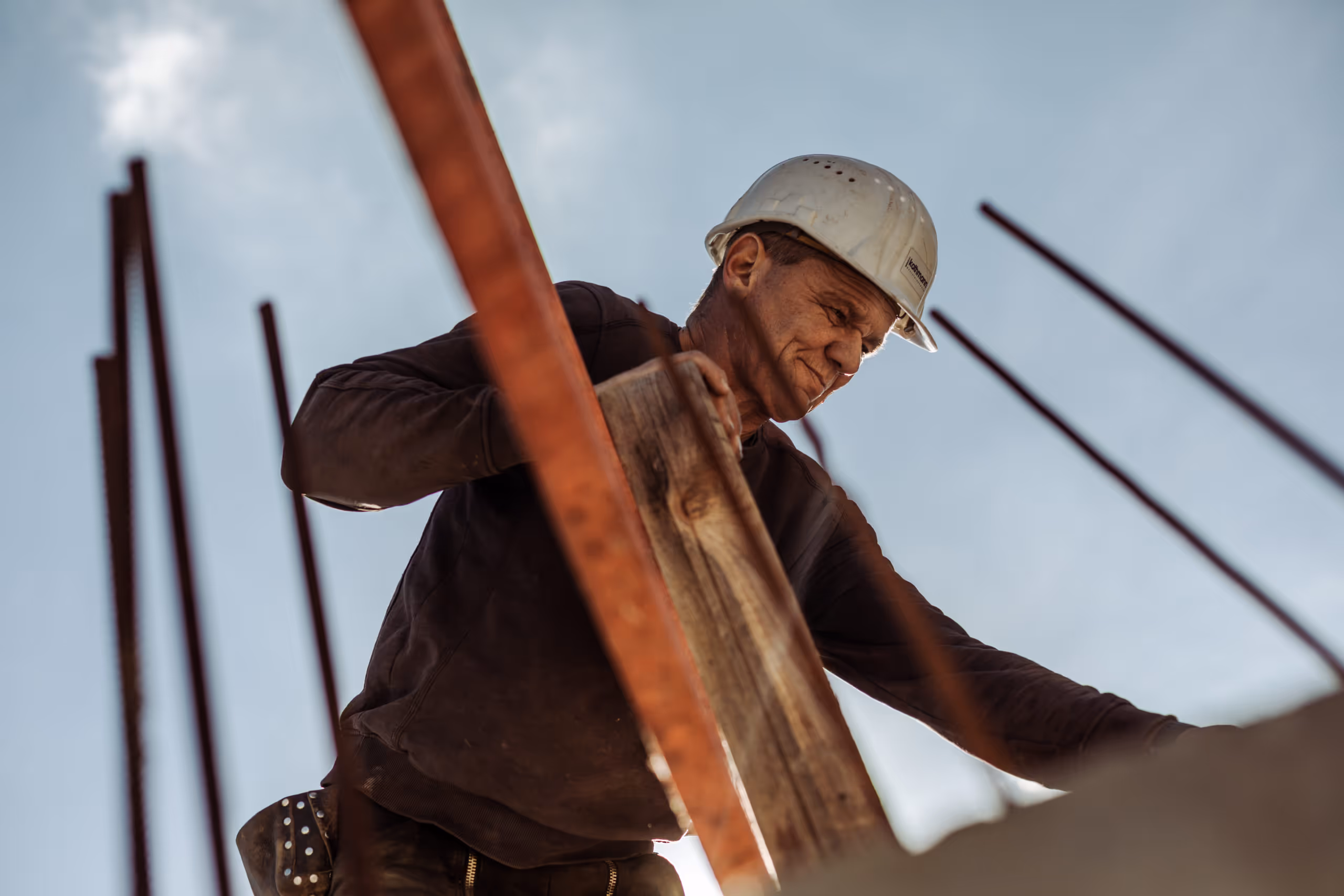 Baustellenarbeiter trägt weißen Schutzhelm und hält Holzlatte mit Metallstäben vor blauem Himmel.