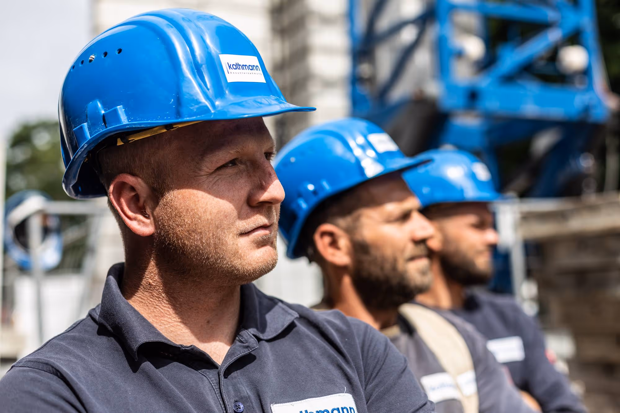 Drei Bauarbeiter mit blauen Schutzhelmen und dunklen Hemden blicken konzentriert nach rechts.