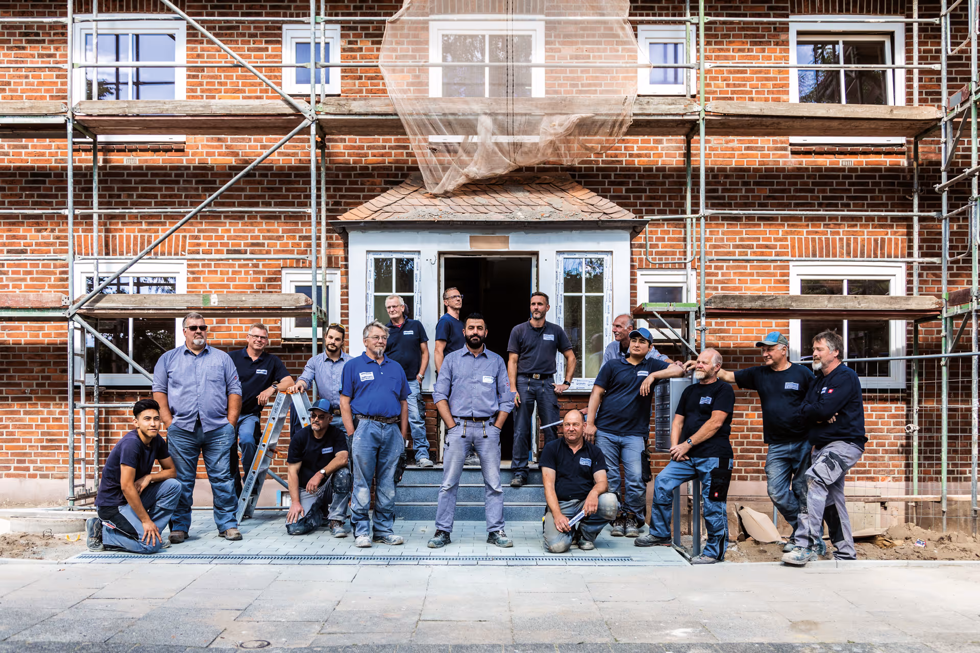 Gruppenfoto von 16 Handwerkern vor einem Backsteingebäude mit Gerüst und einer Eingangstür.