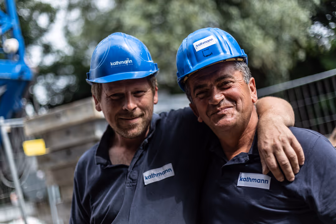 Zwei Bauarbeiter mit blauen Helmen und hellblauen Hemden umarmen sich und lächeln auf einer Baustelle.