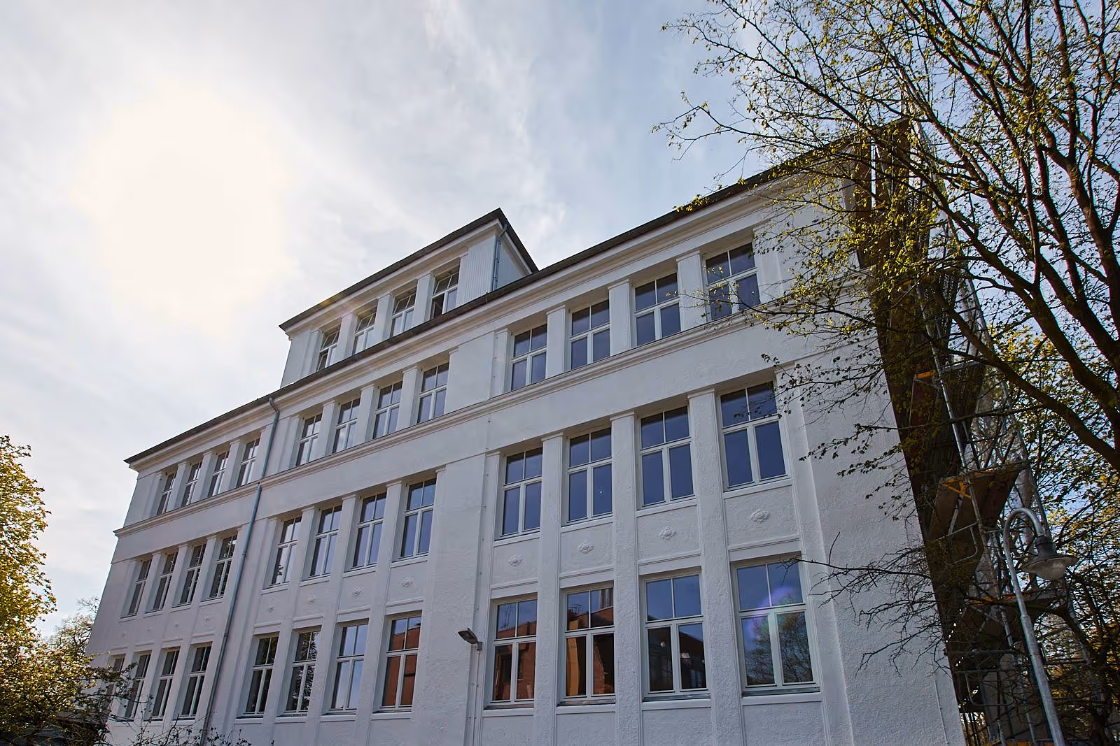 Weißes mehrstöckiges Gebäude mit vielen Fenstern und Bäumen im Vordergrund bei bewölktem Himmel.