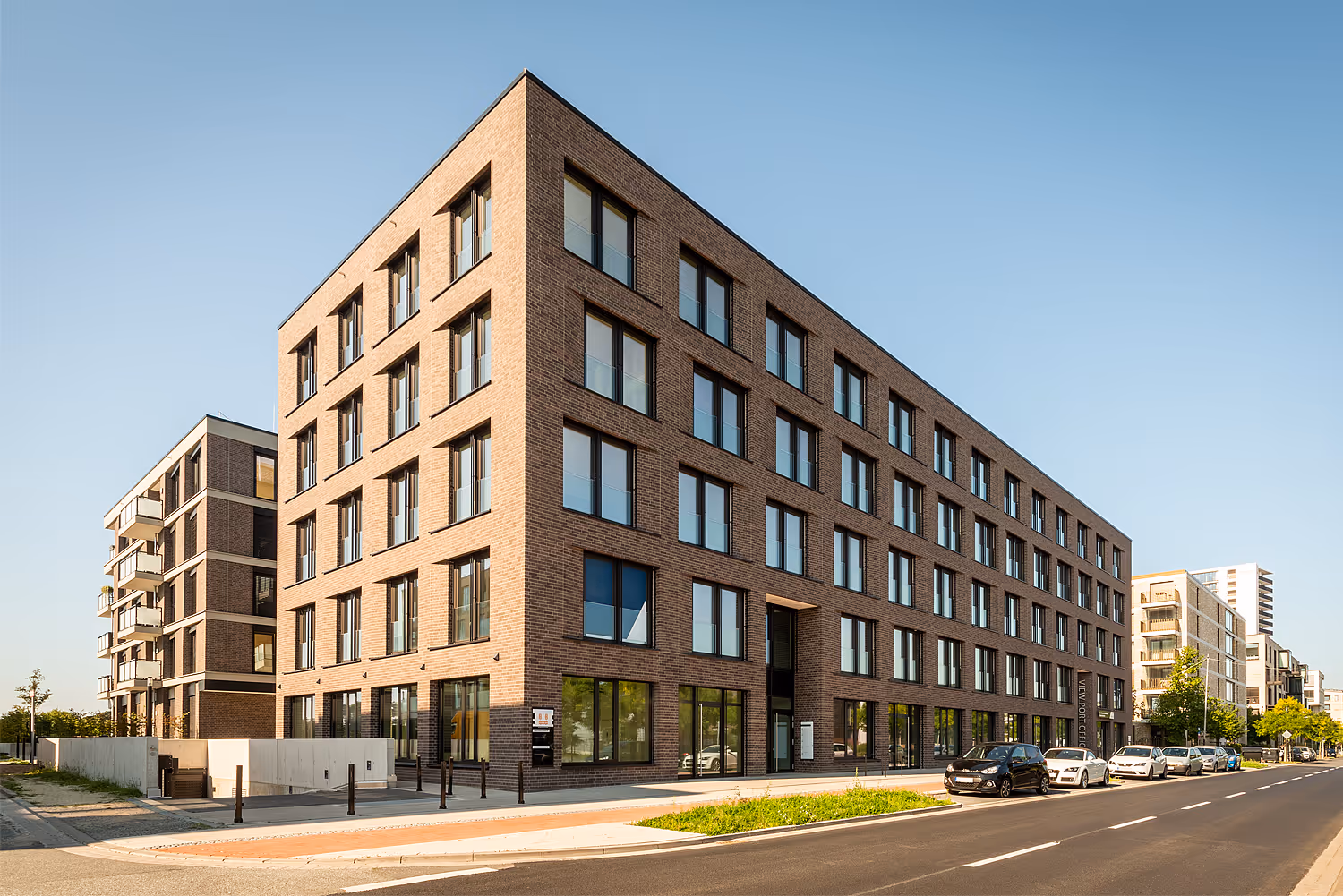 Modernes mehrstöckiges Bürogebäude aus Backstein an einer sonnigen Straße mit parkenden Autos.