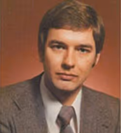 Porträt eines Mannes mit braunen Haaren, der einen grau karierten Anzug und Krawatte trägt vor einem braunen Hintergrund.